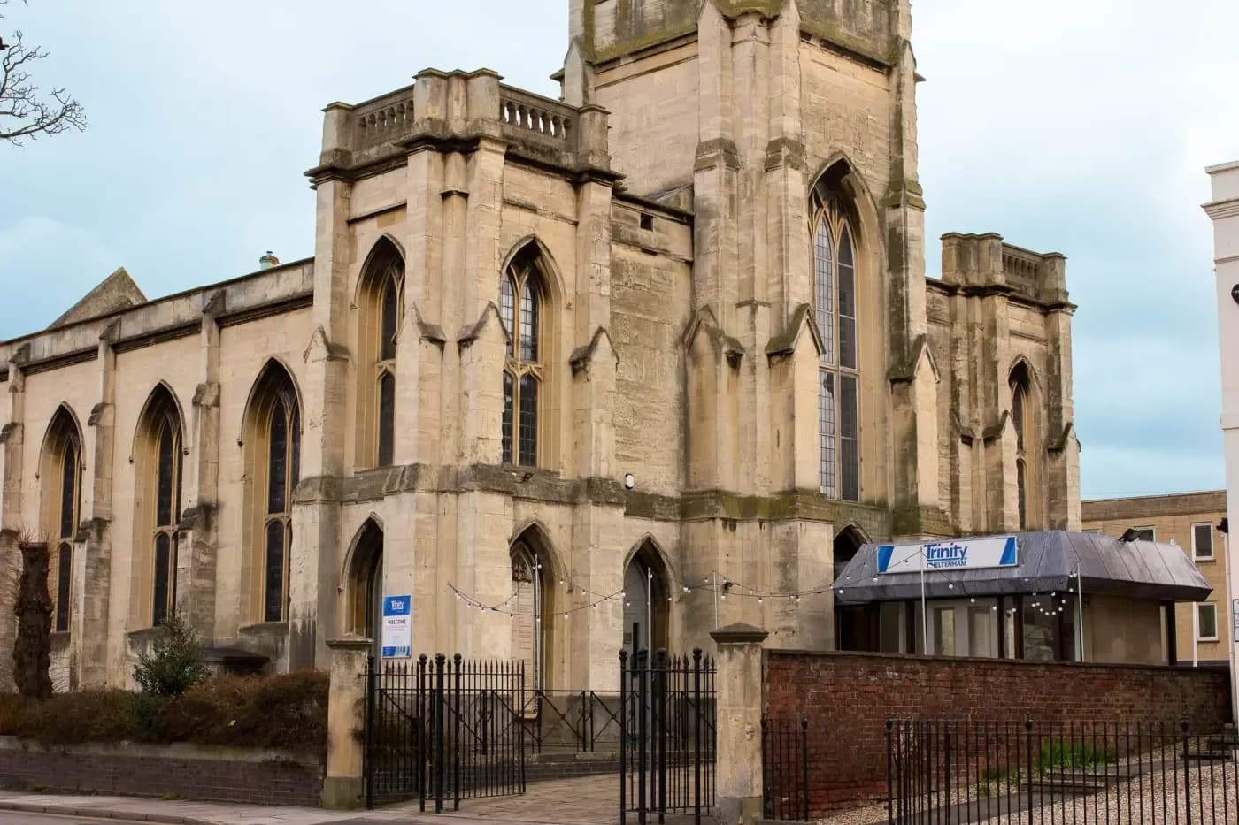 Exterior of Trinity Cheltenham, a historic stone church with arched windows and a tower, with a welcome sign at the entrance