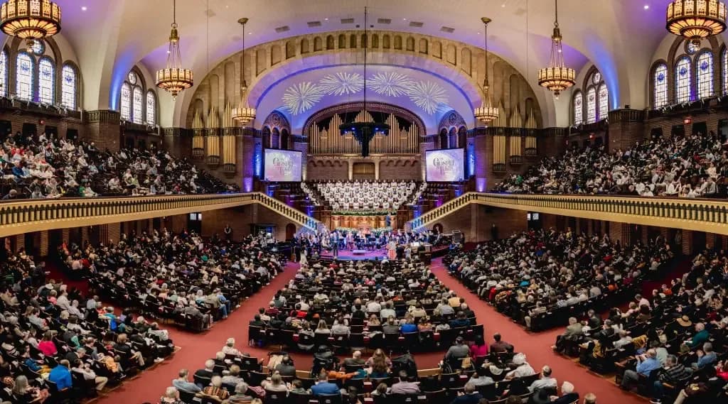 Interior of The Moody Church sanctuary filled with a large congregation, featuring the ornate domed ceiling, chandeliers, stained-glass windows, pipe organ, and choir during a worship service