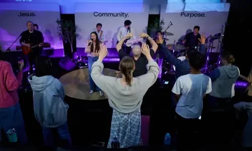 Congregation worshipping with raised hands during a C3 SYD service, with the band on stage and 'Jesus. Community. Purpose.' displayed behind